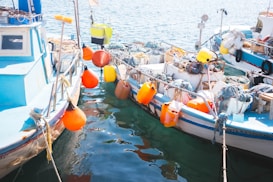Two small fishing boats are moored side by side in a sunlit harbor. The boats are equipped with colorful fishing gear, including orange and yellow buoys, nets, and ropes. The water is clear, reflecting the light, and various items are scattered across the decks.