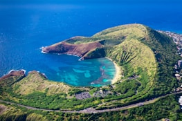 An aerial view of a picturesque bay with turquoise water, surrounded by lush, green hills. The coastline forms a curved, crescent-shaped beach with clear water and visible coral reefs. There are small urban developments near the shore.