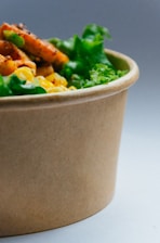 A close-up of a paper bowl filled with a vibrant salad. Visible ingredients include fresh green lettuce, bright yellow corn, and grilled orange carrots. The bowl is set against a simple gray background.