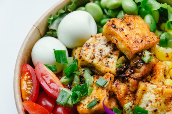 A colorful salad bowl filled with a variety of ingredients including grilled tofu, sliced cherry tomatoes, boiled eggs, edamame, corn, and fresh greens. The tofu is seasoned and appears to have been grilled or roasted, providing a crispy texture.