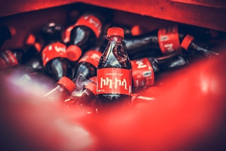 A collection of soda bottles with bold red labels and caps piled together. One bottle is in clear focus at the center, displaying text in a non-Latin script. The color red dominates the scene, and the bottles contrast starkly with the darker background.