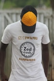 A person wearing a white t-shirt with the text 'Zed Cash' printed on it. The person is also wearing a cap with a distinctive orange quilted pattern on the front panel. The background appears to be outdoors, with a blurred view of a fence or similar structure.