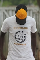 A person wearing a white t-shirt with the text 'Zed Cash' printed on it. The person is also wearing a cap with a distinctive orange quilted pattern on the front panel. The background appears to be outdoors, with a blurred view of a fence or similar structure.