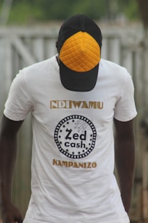A person wearing a white t-shirt with the text 'Zed Cash' printed on it. The person is also wearing a cap with a distinctive orange quilted pattern on the front panel. The background appears to be outdoors, with a blurred view of a fence or similar structure.