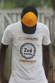 A person wearing a white t-shirt with the text 'Zed Cash' printed on it. The person is also wearing a cap with a distinctive orange quilted pattern on the front panel. The background appears to be outdoors, with a blurred view of a fence or similar structure.