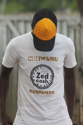 A person wearing a white t-shirt with the text 'Zed Cash' printed on it. The person is also wearing a cap with a distinctive orange quilted pattern on the front panel. The background appears to be outdoors, with a blurred view of a fence or similar structure.