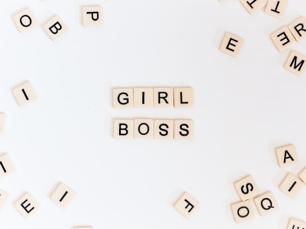A collection of letter tiles on a white surface, arranged to form the words 'GIRL BOSS' in the center, surrounded by scattered, unarranged tiles.
