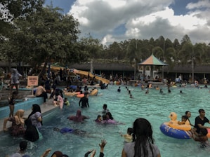 A crowded outdoor swimming pool filled with people of various ages enjoying the water. There is a water slide in the background and several trees and palm trees bordering the pool area. Some people are lounging around the edges while children and adults swim and play. A few inflatable toys, including one shaped like a duck, are in the water. Overcast skies provide a shaded atmosphere.