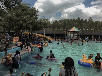 A crowded outdoor swimming pool filled with people of various ages enjoying the water. There is a water slide in the background and several trees and palm trees bordering the pool area. Some people are lounging around the edges while children and adults swim and play. A few inflatable toys, including one shaped like a duck, are in the water. Overcast skies provide a shaded atmosphere.
