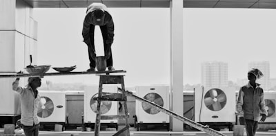 Three construction workers are engaged in a task at a building site. One person is standing on a ladder, focused on balancing a plank while another worker stands on the plank, bending over to adjust something. A third worker stands nearby, observing. The scene unfolds against the backdrop of large industrial fans and distant buildings, creating an urban construction atmosphere.
