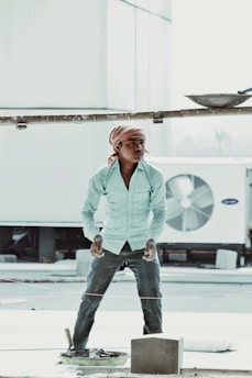 A person wearing a striped long-sleeve shirt, dark trousers, and a headscarf stands in an outdoor setting, appearing to be involved in construction work. The person is positioned next to a large industrial air conditioning unit and construction materials are visible, including a cement block and a trowel.