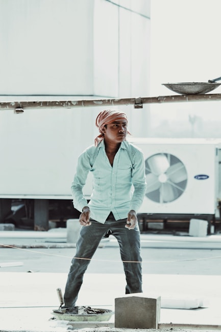 A person wearing a striped long-sleeve shirt, dark trousers, and a headscarf stands in an outdoor setting, appearing to be involved in construction work. The person is positioned next to a large industrial air conditioning unit and construction materials are visible, including a cement block and a trowel.
