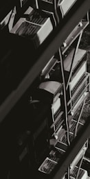 A monochromatic image showcasing a series of industrial air conditioning units stacked vertically against the side of a building. The units have a grid-like design on top, and metal railings can be seen alongside them, creating geometric patterns of light and shadow.