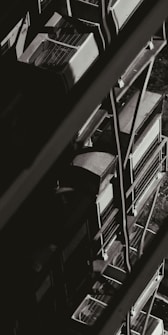 A monochromatic image showcasing a series of industrial air conditioning units stacked vertically against the side of a building. The units have a grid-like design on top, and metal railings can be seen alongside them, creating geometric patterns of light and shadow.