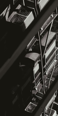 A monochromatic image showcasing a series of industrial air conditioning units stacked vertically against the side of a building. The units have a grid-like design on top, and metal railings can be seen alongside them, creating geometric patterns of light and shadow.