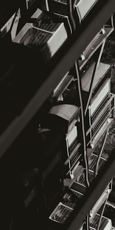 A monochromatic image showcasing a series of industrial air conditioning units stacked vertically against the side of a building. The units have a grid-like design on top, and metal railings can be seen alongside them, creating geometric patterns of light and shadow.