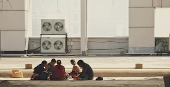 A group of people is seated in a circle on the ground inside a large, open, industrial space. The background features a wall with several air conditioning units mounted on it. Sunlight casts strong shadows, suggesting an incomplete structure or under-construction building.