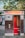 A vintage photo booth with a sign that reads 'Photoautomat', featuring a wooden exterior and red interior. The booth is positioned outdoors with trees and a building in the background. Posters and graffiti are visible on the adjacent wall.