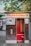 A vintage photo booth with a sign that reads 'Photoautomat', featuring a wooden exterior and red interior. The booth is positioned outdoors with trees and a building in the background. Posters and graffiti are visible on the adjacent wall.