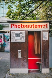 A vintage photo booth with a sign that reads 'Photoautomat', featuring a wooden exterior and red interior. The booth is positioned outdoors with trees and a building in the background. Posters and graffiti are visible on the adjacent wall.