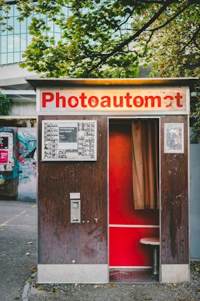 A vintage photo booth with a sign that reads 'Photoautomat', featuring a wooden exterior and red interior. The booth is positioned outdoors with trees and a building in the background. Posters and graffiti are visible on the adjacent wall.