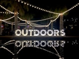 Large, illuminated letters spelling 'OUTDOORS' are displayed in a nighttime setting. String lights and palm trees add to the festive ambiance. Reflections of the lights and letters are visible on a nearby water surface.