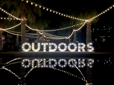 Large, illuminated letters spelling 'OUTDOORS' are displayed in a nighttime setting. String lights and palm trees add to the festive ambiance. Reflections of the lights and letters are visible on a nearby water surface.