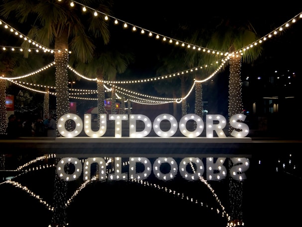 Large, illuminated letters spelling 'OUTDOORS' are displayed in a nighttime setting. String lights and palm trees add to the festive ambiance. Reflections of the lights and letters are visible on a nearby water surface.