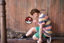 A young person is kneeling on a wooden floor against a graffiti-covered wall, pointing at a pixelated image of a Poké Ball. The individual is wearing a colorful striped shirt, green shorts, and casual shoes. A vertical post is visible on the left.