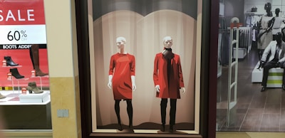 A retail store display features a large poster with two stylized drawings of mannequins. One wears a bright red dress with black leggings and the other sports a red coat over a black outfit. To the left, there's a signage indicating a sale offering up to 60% off on boots, with several shoe displays below it. On the right, inside the store, more mannequins showcase clothing, with a suited mannequin seated on a bench.