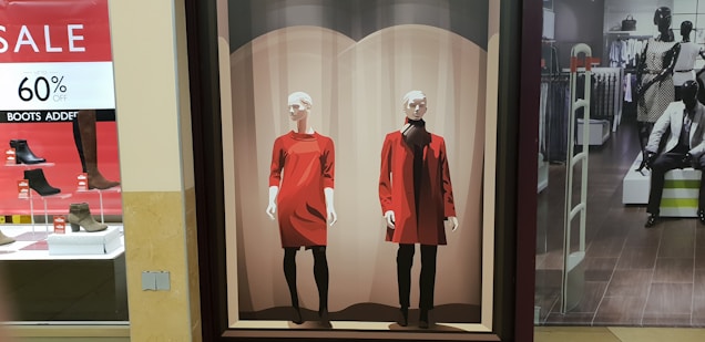 A retail store display features a large poster with two stylized drawings of mannequins. One wears a bright red dress with black leggings and the other sports a red coat over a black outfit. To the left, there's a signage indicating a sale offering up to 60% off on boots, with several shoe displays below it. On the right, inside the store, more mannequins showcase clothing, with a suited mannequin seated on a bench.