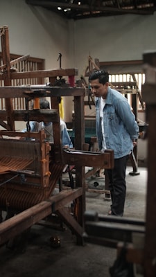 A person is observing a traditional wooden loom in a workshop setting. The loom has various spools of thread, and another person is partially visible, possibly operating the loom. The interior of the space is rustic, with wooden beams and muted lighting.