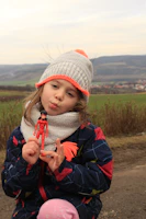 A young child, wearing a knitted hat with a pom-pom and a scarf, poses outdoors with a small toy figure in hand. The child is dressed in a colorful winter coat with a playful expression. The background features a scenic rural landscape with green fields and distant hills.