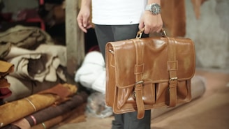 A person is holding a tan leather briefcase, with a watch visible on their wrist. The background includes rolled and stacked pieces of fabric in a storage area, suggesting a workshop or leather store.