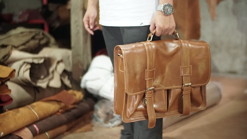 A person is holding a tan leather briefcase, with a watch visible on their wrist. The background includes rolled and stacked pieces of fabric in a storage area, suggesting a workshop or leather store.