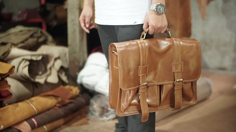 A person is holding a tan leather briefcase, with a watch visible on their wrist. The background includes rolled and stacked pieces of fabric in a storage area, suggesting a workshop or leather store.