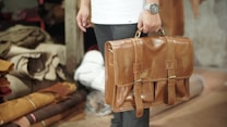 A person is holding a tan leather briefcase, with a watch visible on their wrist. The background includes rolled and stacked pieces of fabric in a storage area, suggesting a workshop or leather store.