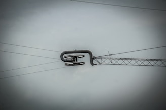 An overhead power line suspension system with metal cables and a structural component, set against a gray sky. The central feature is a curved metal rod supporting several insulators.
