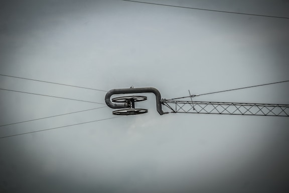 An overhead power line suspension system with metal cables and a structural component, set against a gray sky. The central feature is a curved metal rod supporting several insulators.