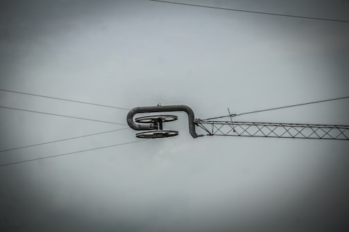 An overhead power line suspension system with metal cables and a structural component, set against a gray sky. The central feature is a curved metal rod supporting several insulators.