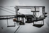 An industrial structure with metal components, wires, and pulleys is prominently featured against a cloudy sky. The assembly includes a rectangular frame with cables and electronic components, suggesting utility or telecommunications equipment.