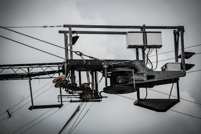 An industrial structure with metal components, wires, and pulleys is prominently featured against a cloudy sky. The assembly includes a rectangular frame with cables and electronic components, suggesting utility or telecommunications equipment.
