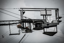 An industrial structure with metal components, wires, and pulleys is prominently featured against a cloudy sky. The assembly includes a rectangular frame with cables and electronic components, suggesting utility or telecommunications equipment.