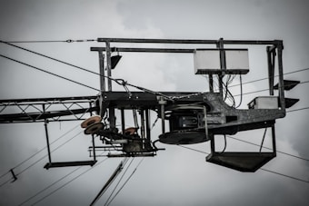 An industrial structure with metal components, wires, and pulleys is prominently featured against a cloudy sky. The assembly includes a rectangular frame with cables and electronic components, suggesting utility or telecommunications equipment.