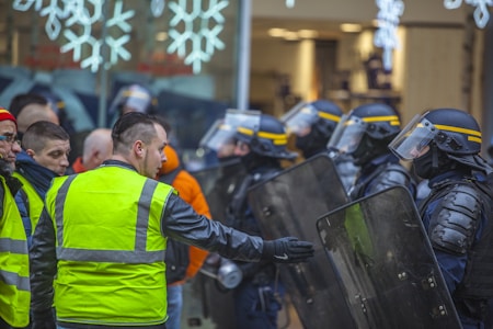 A group of people wearing yellow vests faces a line of riot police equipped with helmets, visors, and shields. The setting appears to be tense, suggesting a protest or demonstration. The police are standing in formation, while the individuals in vests seem to be engaging with them.