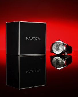 A sophisticated watch with a silver casing and black leather strap is positioned beside a black Nautica watch box. The background and reflection surface are bright red, creating a dramatic contrast.