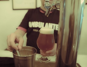 A person wearing a dark shirt with visible text is holding a glass of beer with a foamy head. Their other hand is seen stirring or holding a utensil in a metal container. A beer tap is prominently in the foreground.
