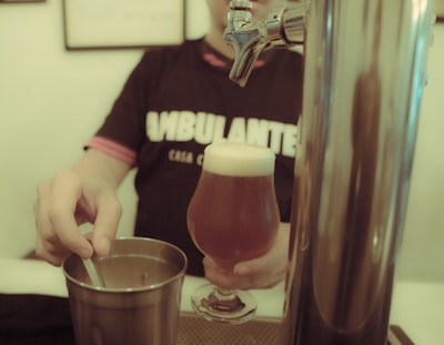 A person wearing a dark shirt with visible text is holding a glass of beer with a foamy head. Their other hand is seen stirring or holding a utensil in a metal container. A beer tap is prominently in the foreground.