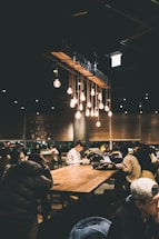 A dimly lit, cozy café filled with people sitting at long wooden tables. Warm hanging light bulbs create a relaxed atmosphere. Several individuals are engaged with their laptops and phones, suggesting a busy yet peaceful environment.