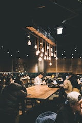 A dimly lit, cozy café filled with people sitting at long wooden tables. Warm hanging light bulbs create a relaxed atmosphere. Several individuals are engaged with their laptops and phones, suggesting a busy yet peaceful environment.